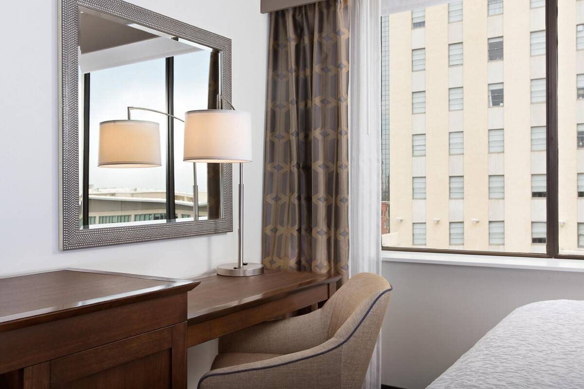 A hotel room with a desk, chair, lamp, and a large window with a view of buildings.