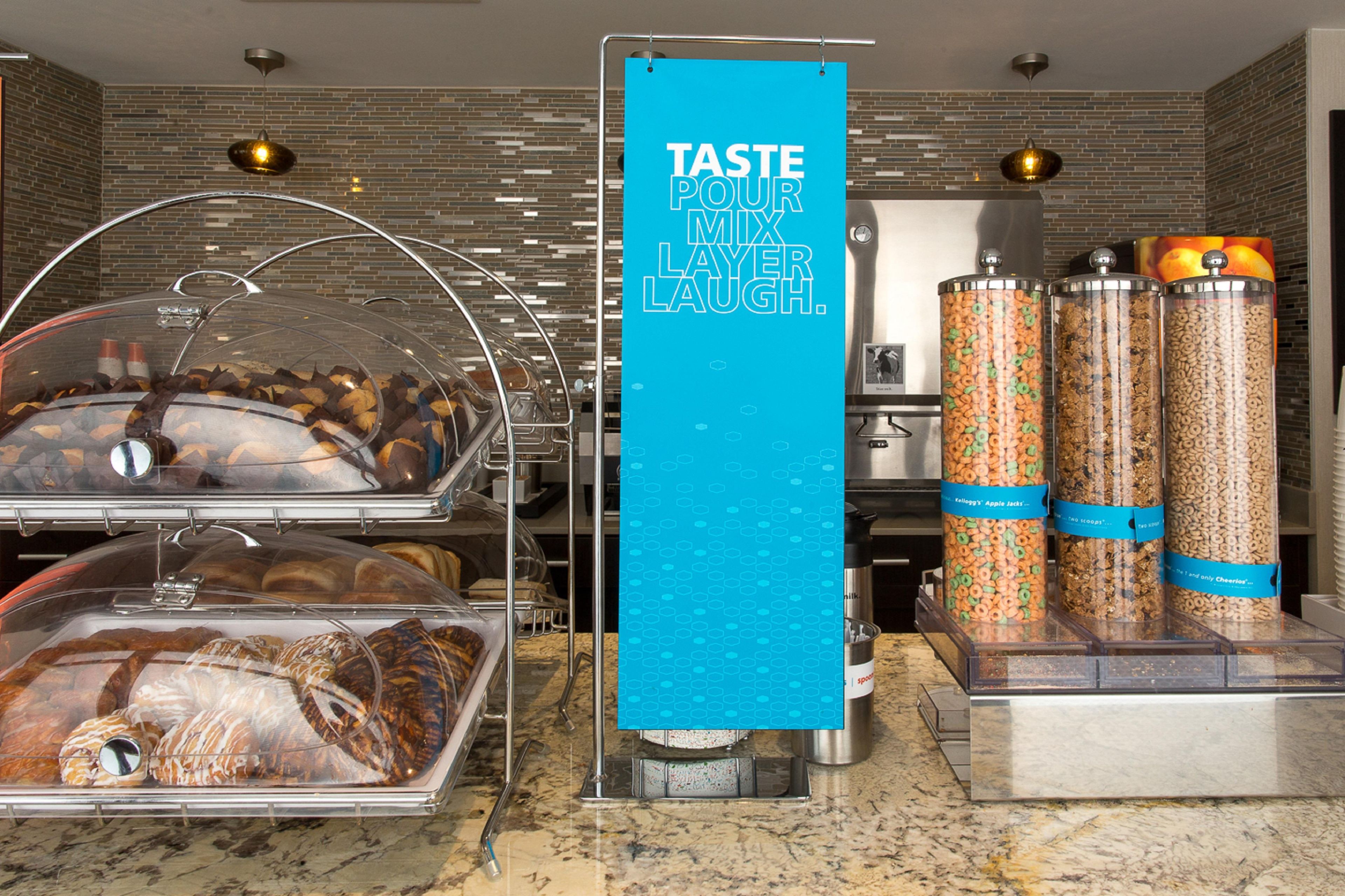 Un espace buffet pour le petit-déjeuner avec une pancarte sur laquelle on peut lire : « GOÛTEZ, PARTAGEZ, RIEZ. » Différents aliments sont présentés dans des récipients transparents, et une machine à café est à disposition.