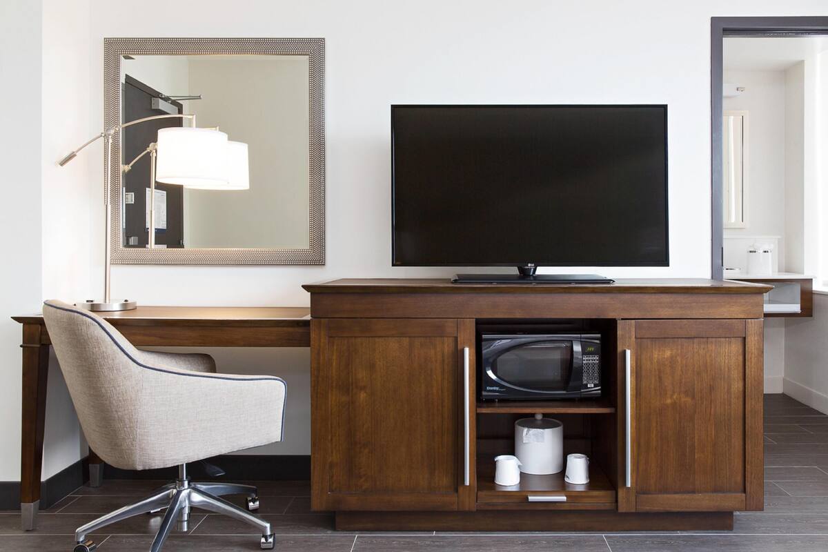 A modern hotel room with a wooden desk, a flat-screen TV, a chair, a microwave, and a mirror.
