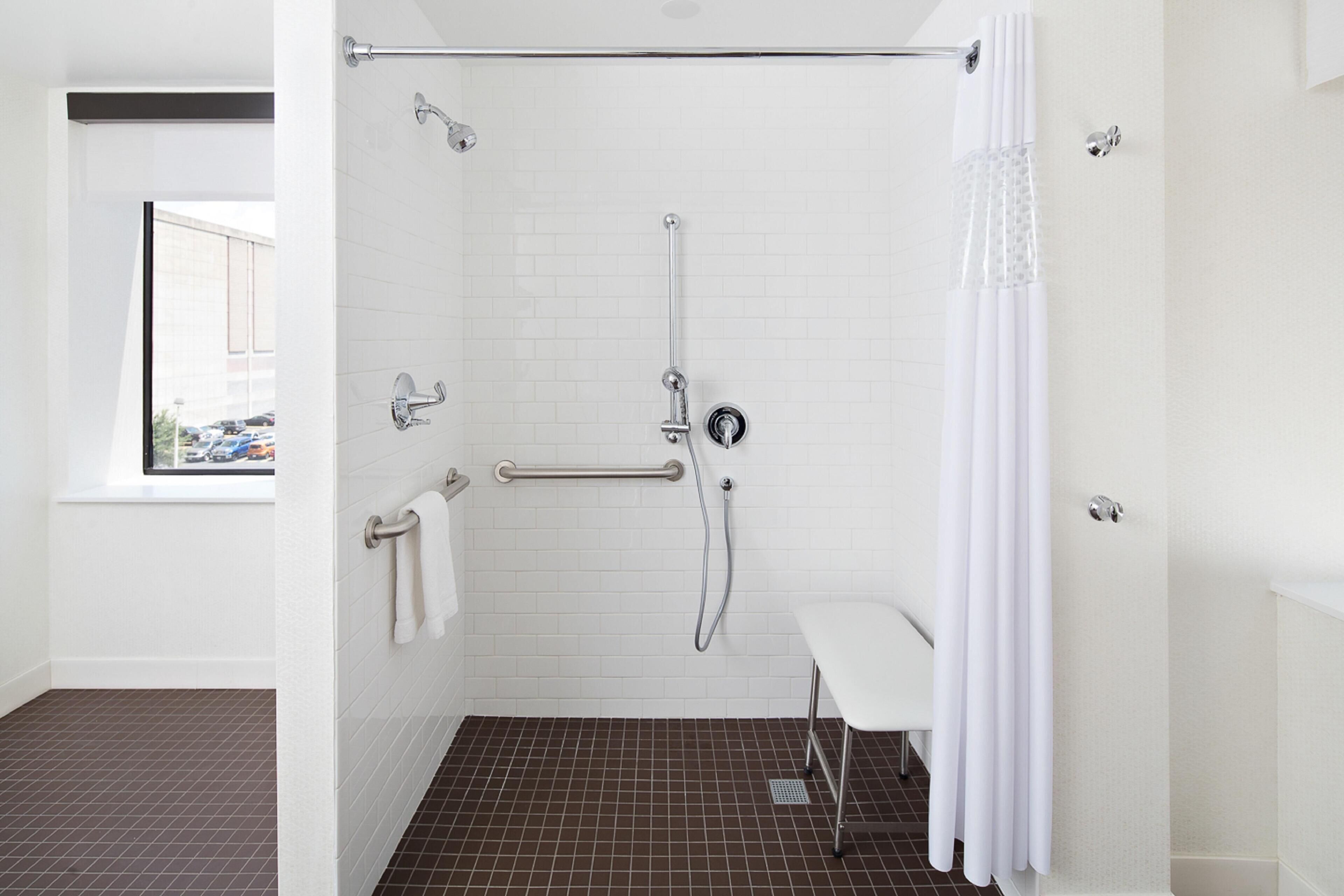 A modern bathroom with white tiled walls, a shower with a grab bar, a white bench, and a window with a view of parked cars.