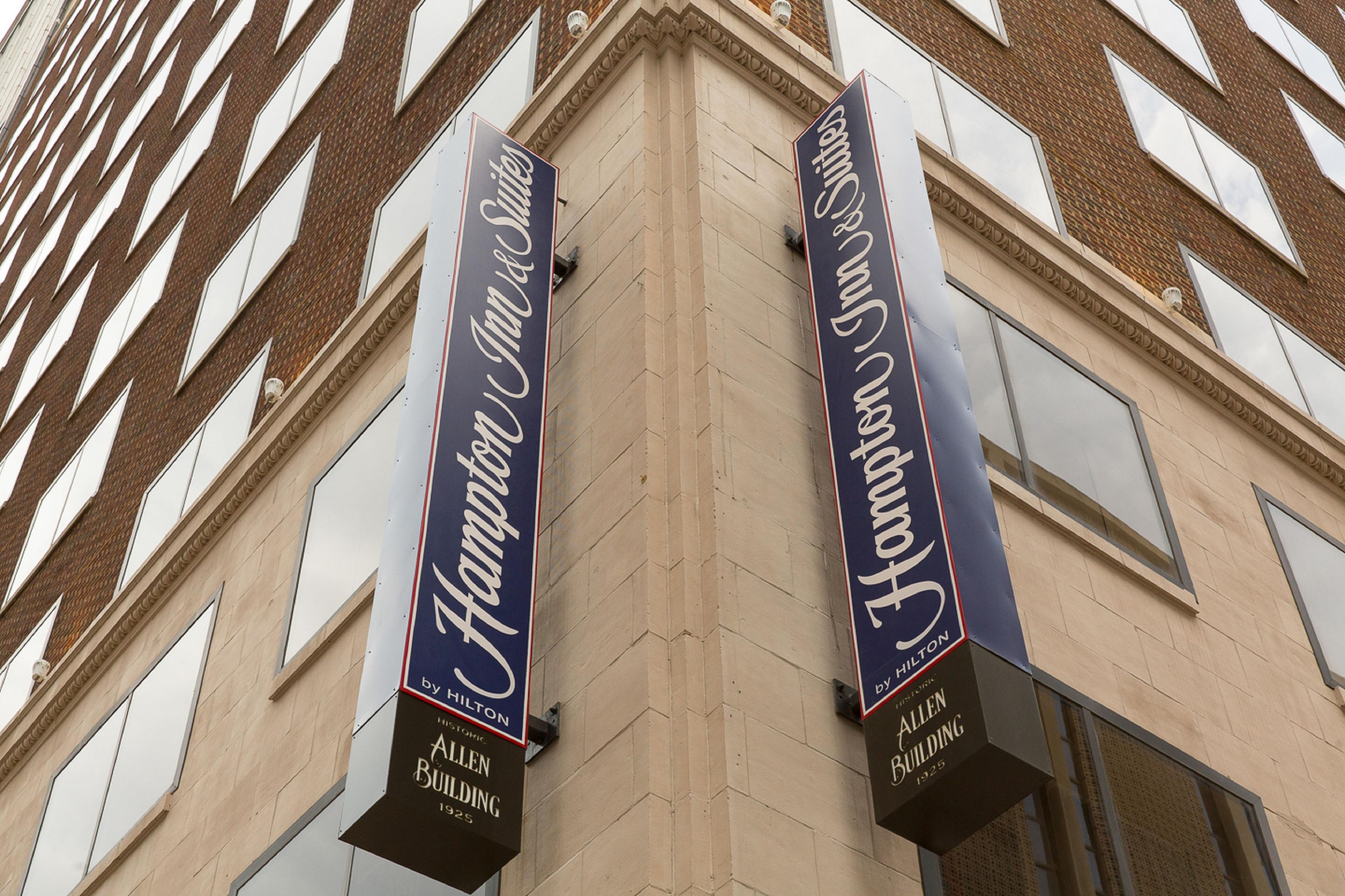 Twee banners voor Hampton Inn Suites by Hilton aan het Allen Building, die de locatie en het lidmaatschap van het hotel aangeven.