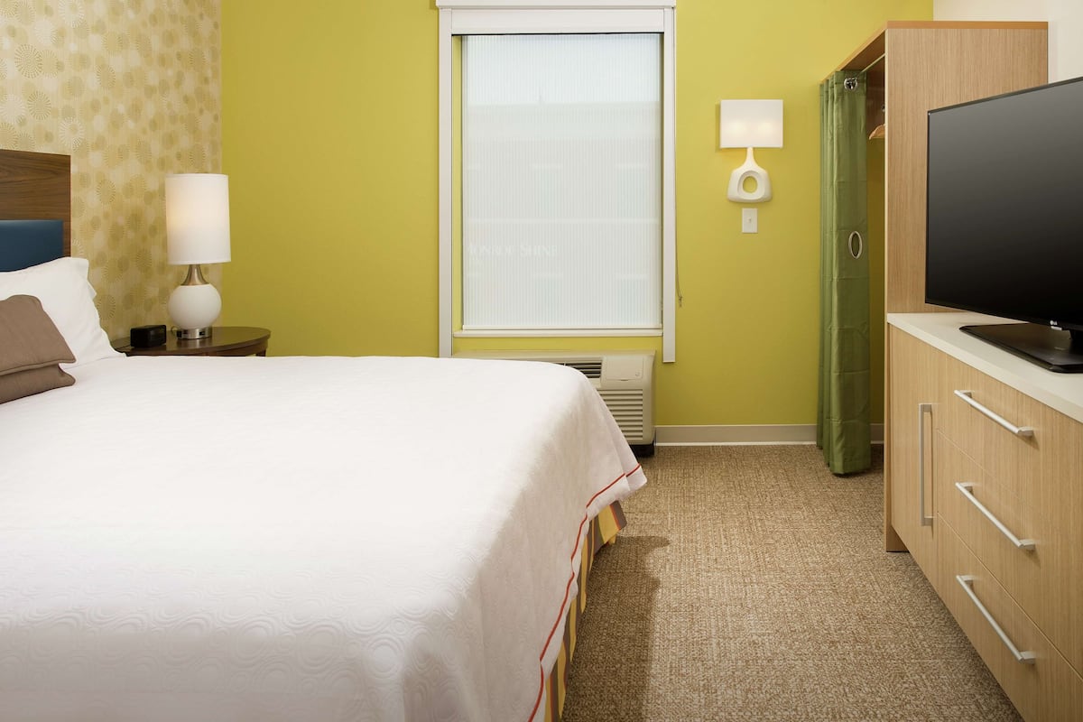 A hotel room with a bed, a bedside lamp, a television, a window with blinds, and a green curtain.