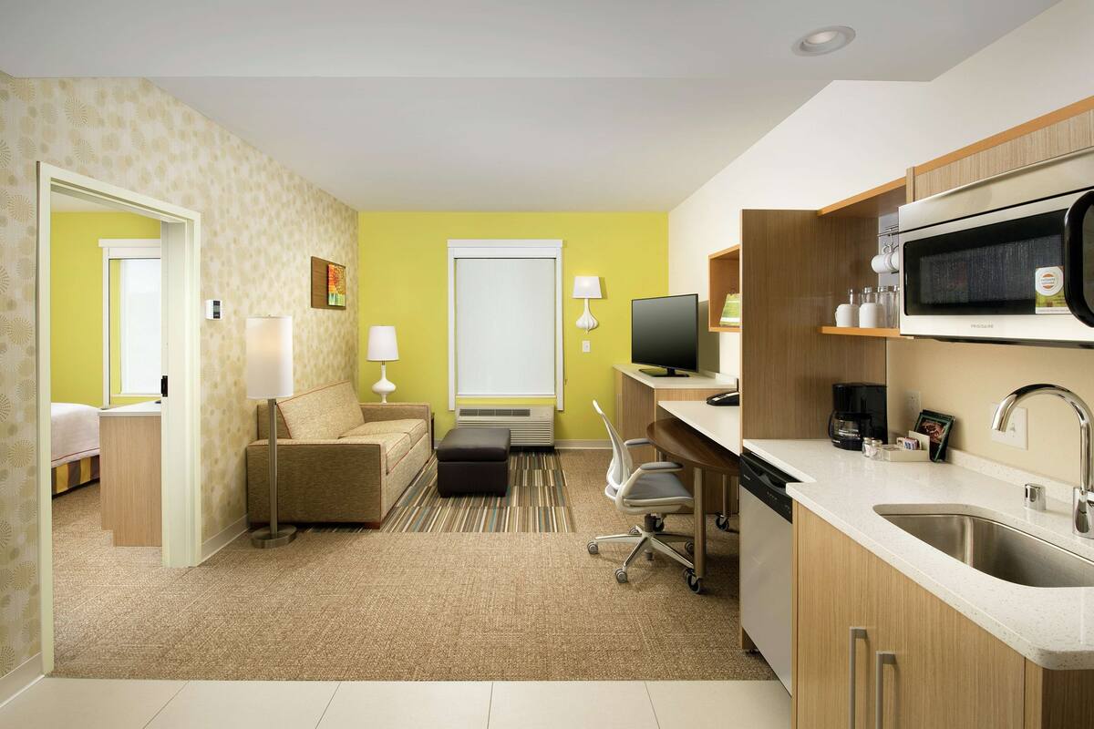 A compact hotel room with a kitchenette, a sofa, a desk, and a television.