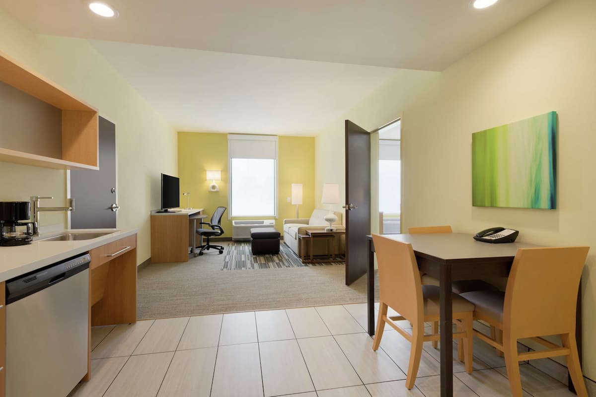 A modern hotel room with a kitchen area, a dining table with chairs, a sofa, a bedside table with a lamp, and a large window.
