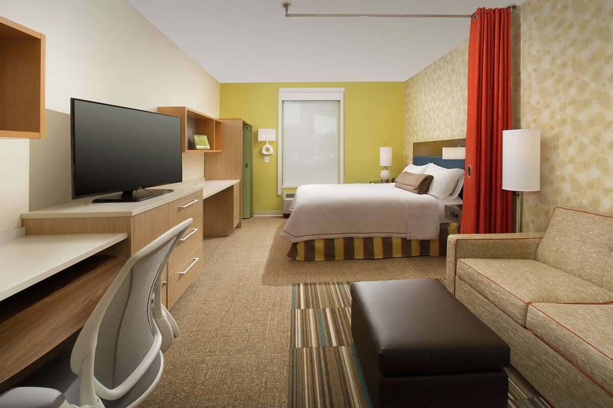 A hotel room with a flat-screen TV, a desk with a chair, a bed with pillows, a sofa, and a red curtain.