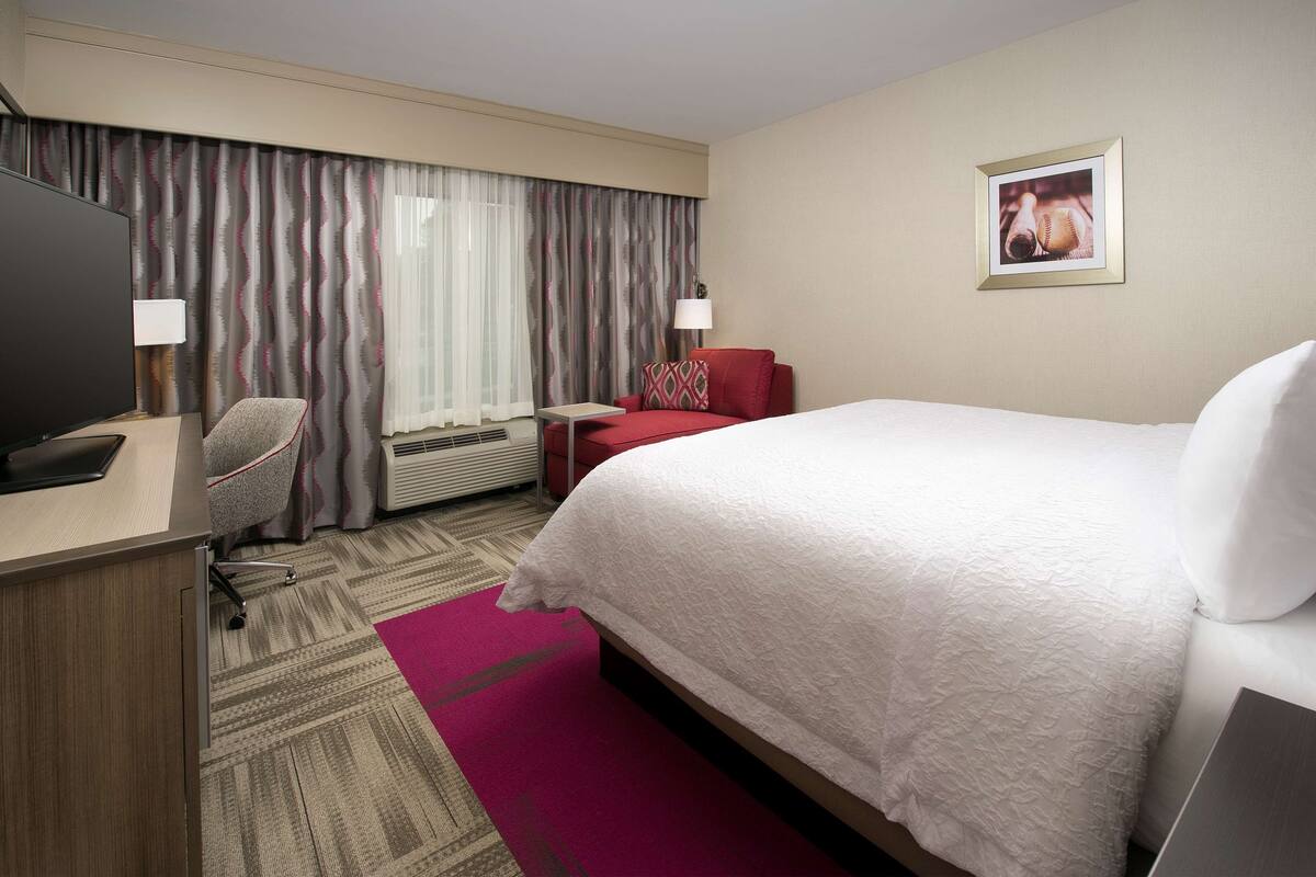A hotel room with a bed, a desk, a chair, a TV, and a window with curtains.