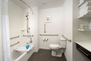 Baño con ducha, inodoro y lavamanos, equipado con barras de apoyo y asiento de ducha accesible para sillas de ruedas.