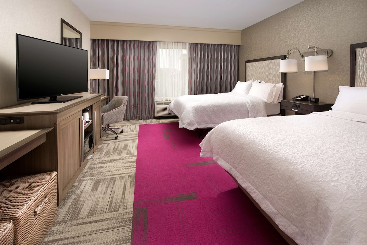A hotel room with a flat-screen TV, two beds, a desk, a chair, and a large window with curtains.