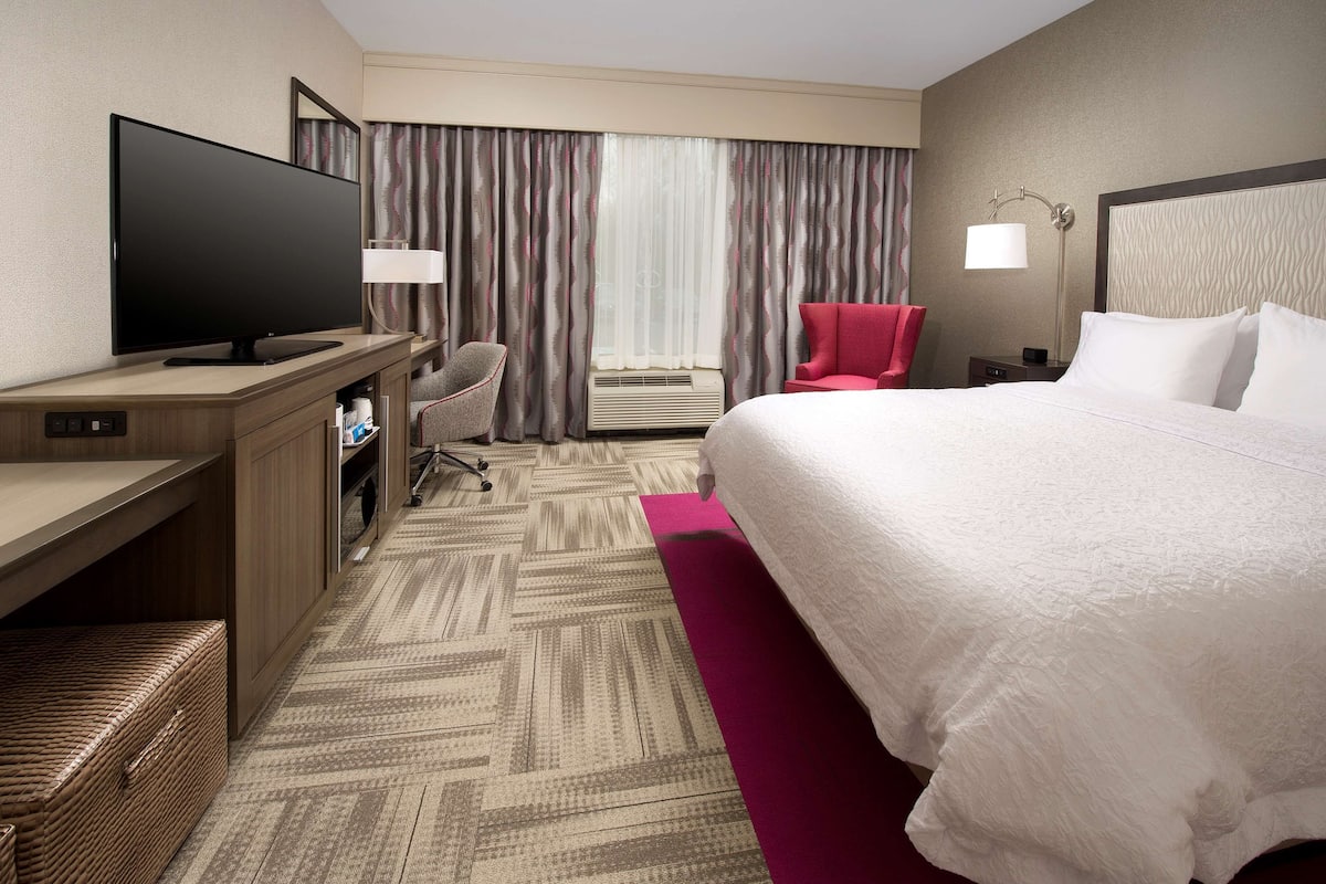 A hotel room with a large bed, a desk with a flat-screen TV, a chair, a nightstand, and a lamp.