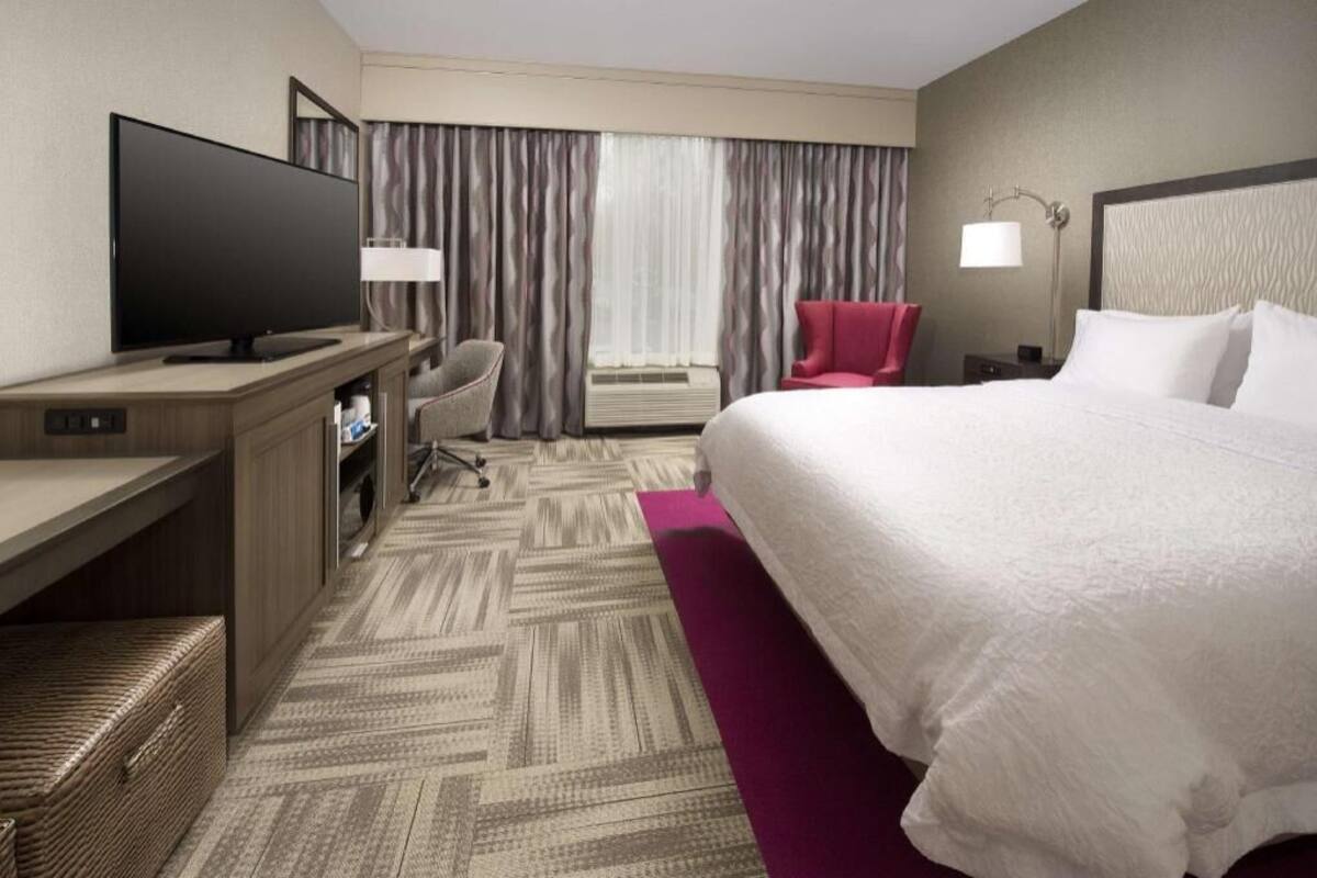 A hotel room with a large bed, a television, a desk with a chair, a red armchair, and a window with curtains.