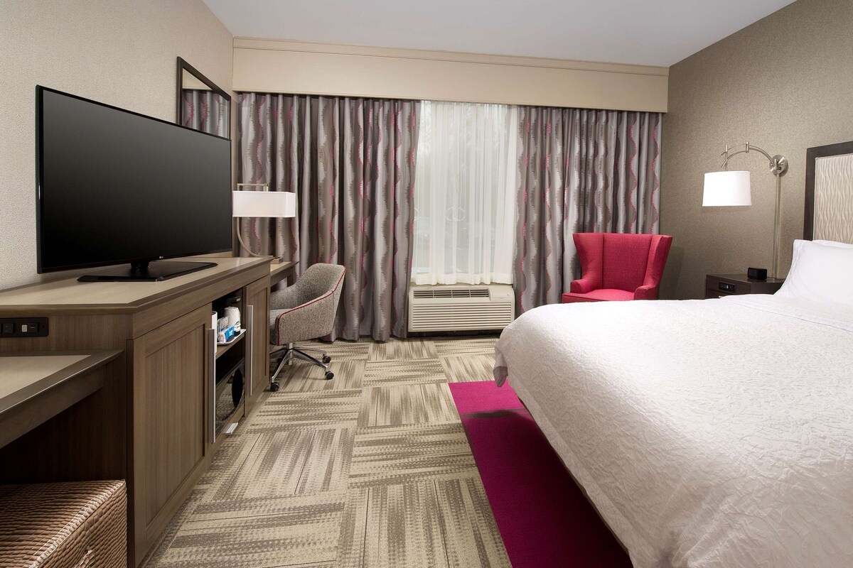 A hotel room with a bed, a desk with a TV, a chair, a window with curtains, and an air conditioning unit.