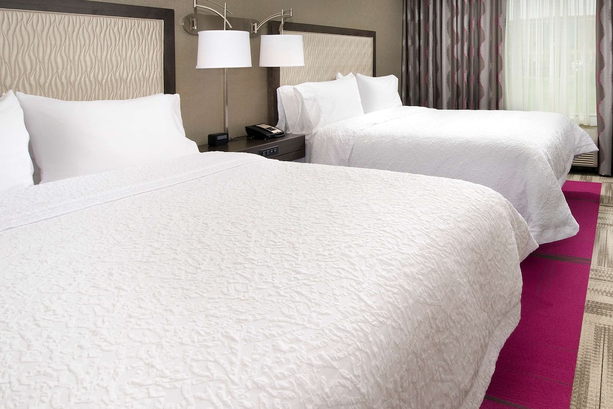 A hotel room with two beds, a headboard, a nightstand with a lamp, and a window with curtains.