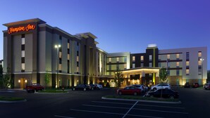 Et Hampton Inn hotel med et moderne design, der har en markant indgang og en parkeringsplads med flere biler.
