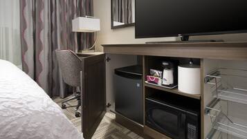 Een hotelkamer met een bed, een bureau, een stoel, een televisie, een prullenbak, een magnetron en een plank met toiletartikelen.