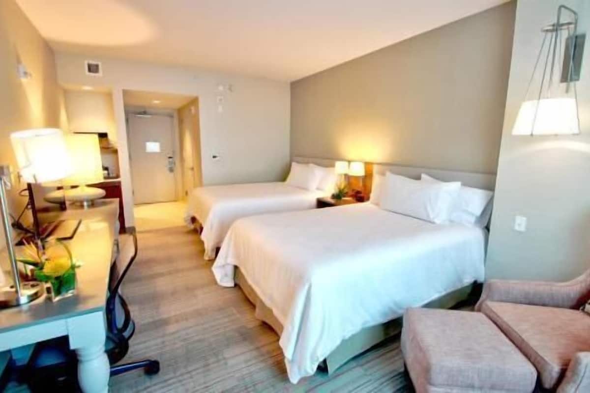 A hotel room with two beds, a desk with a lamp, a chair, and a small table with a vase of flowers.