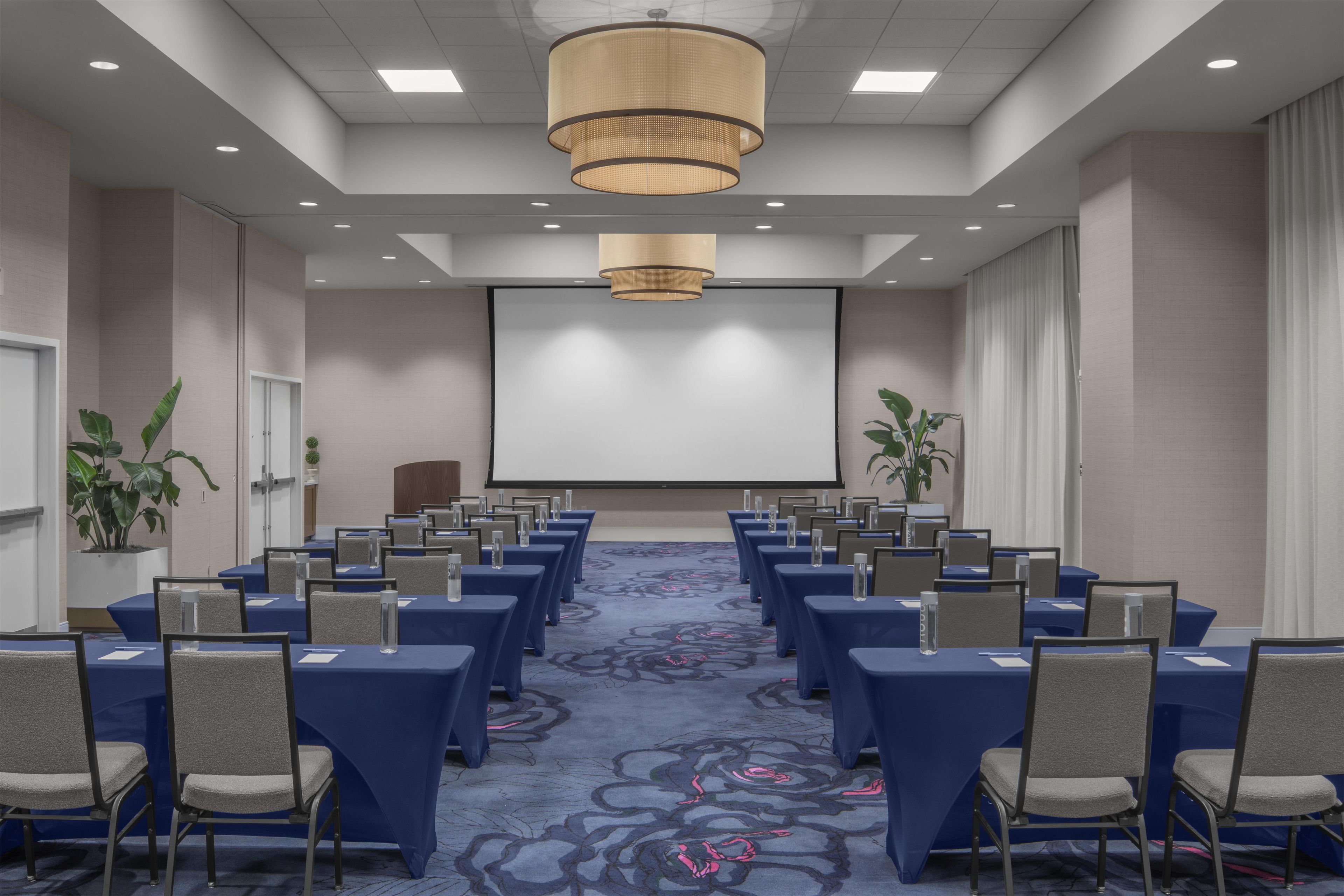 A conference room with rows of tables and chairs, a large screen, and a chandelier.