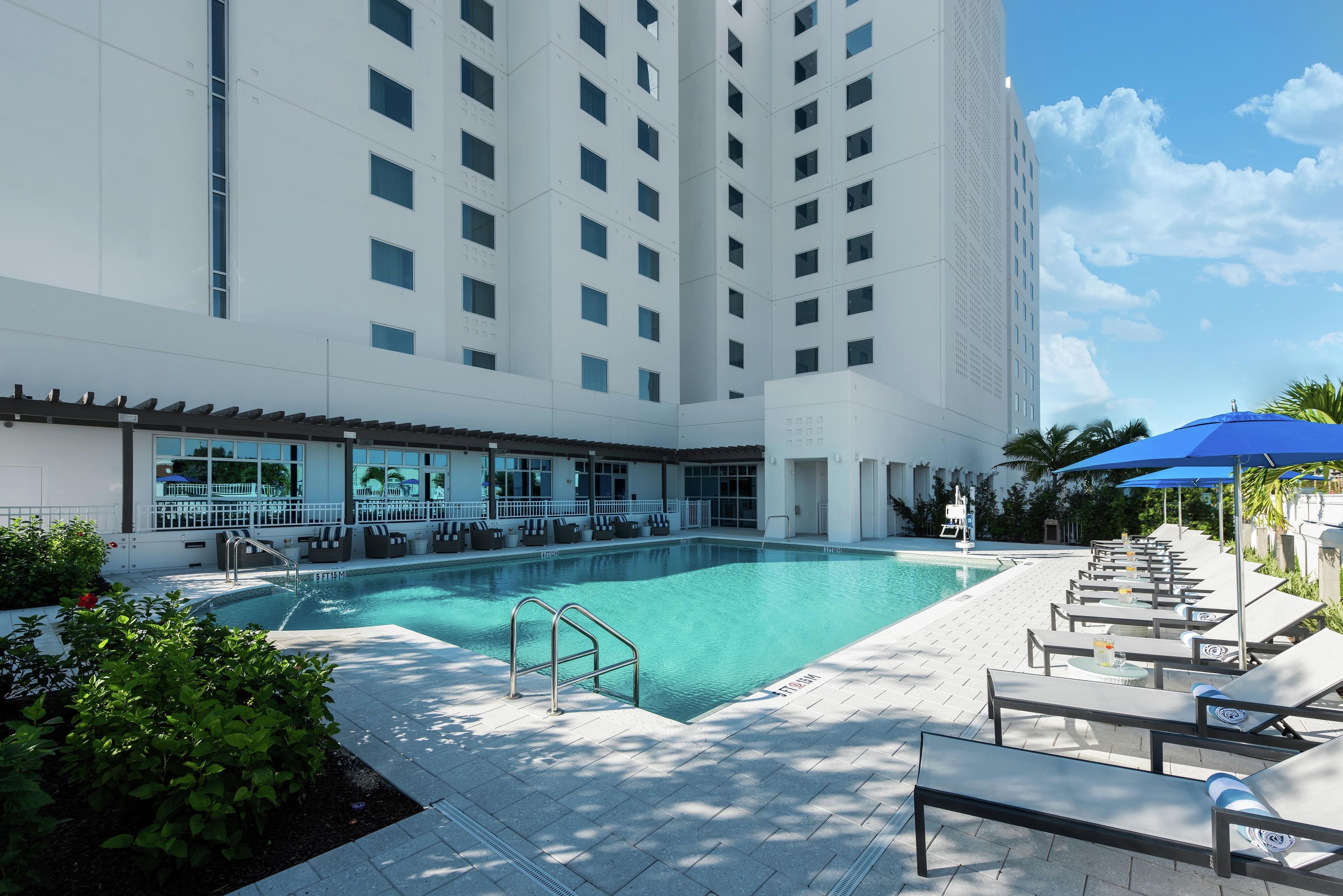 A hotel pool area with a clear pool, sun loungers, and a building with multiple floors in the background.