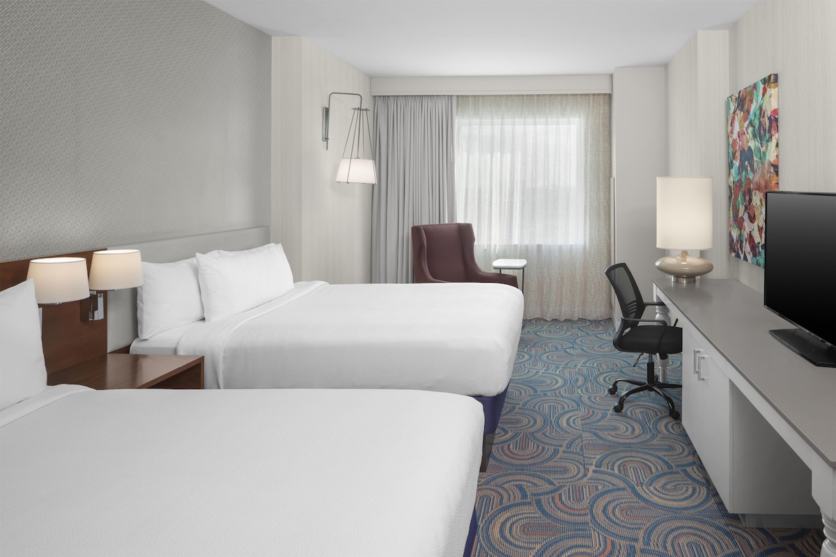 Une chambre d’hôtel avec deux lits, un bureau avec une télévision, une chaise, une lampe et une fenêtre avec des rideaux.