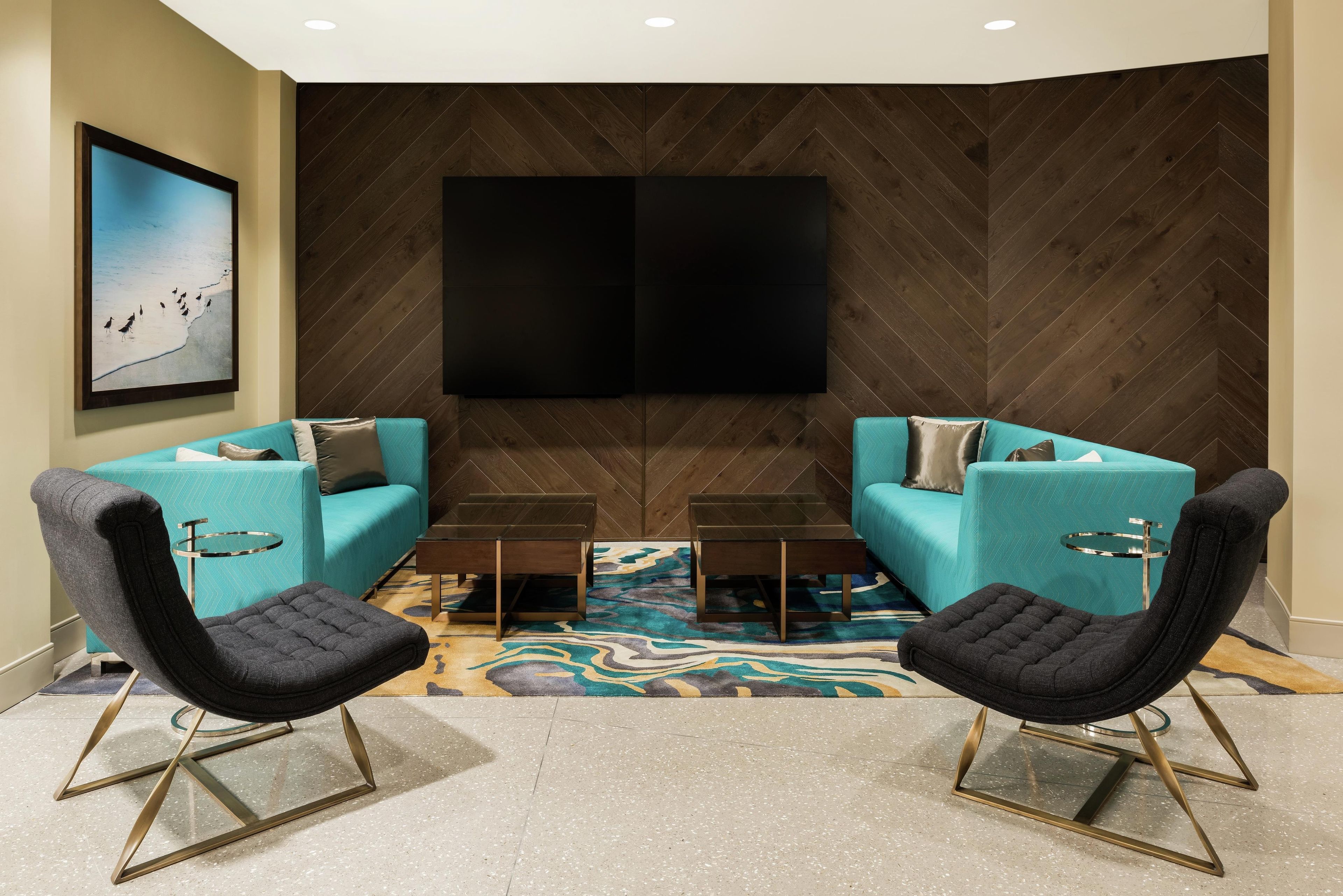 A modern living room with two teal sofas, black chairs, a coffee table, and a large flat-screen TV mounted on a herringbone-patterned wall.