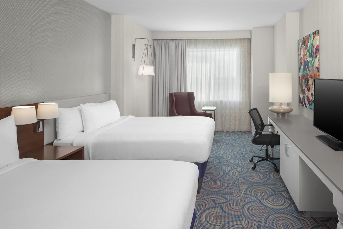 A hotel room with two beds, a desk with a TV, a chair, a lamp, and a window with curtains.