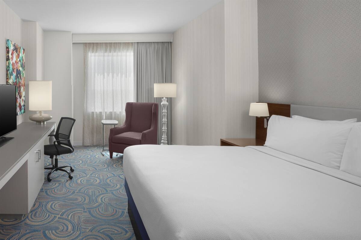A hotel room with a bed, desk, chair, lamps, and a television.