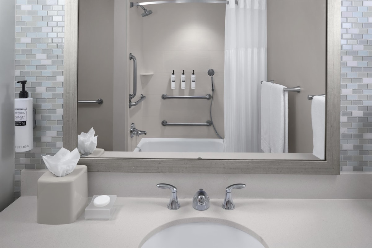 Une salle de bain dotée d’un grand miroir, d’un lavabo à deux robinets, d’un porte-serviettes et d’un espace douche avec une porte en verre.
