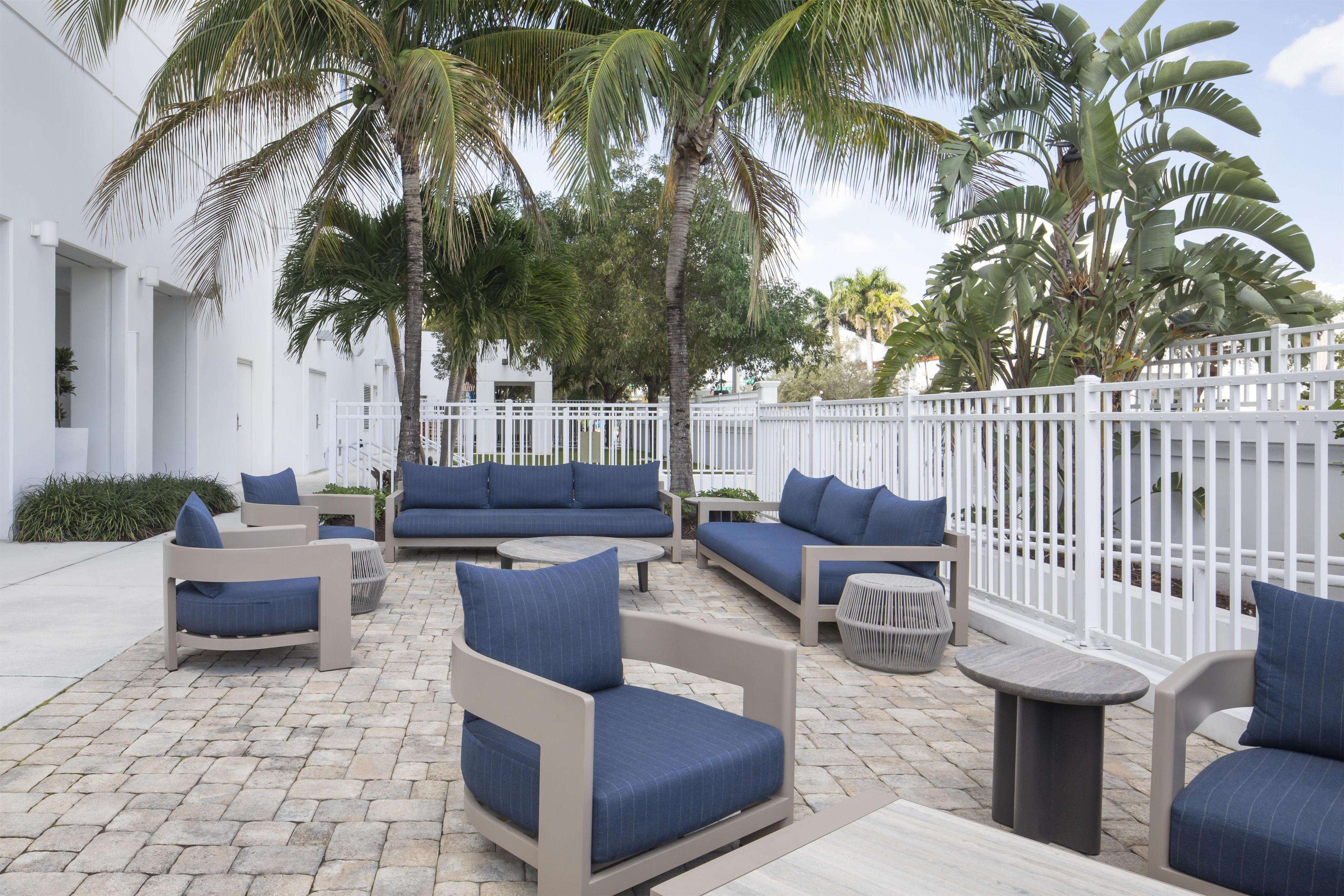 An outdoor seating area with sofas and chairs, palm trees, and a white fence.
