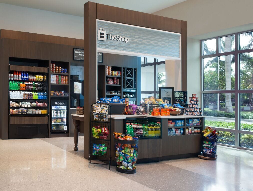 Una tienda de snacks con gran variedad de productos, un refrigerador y vistas al exterior a través de amplios ventanales.