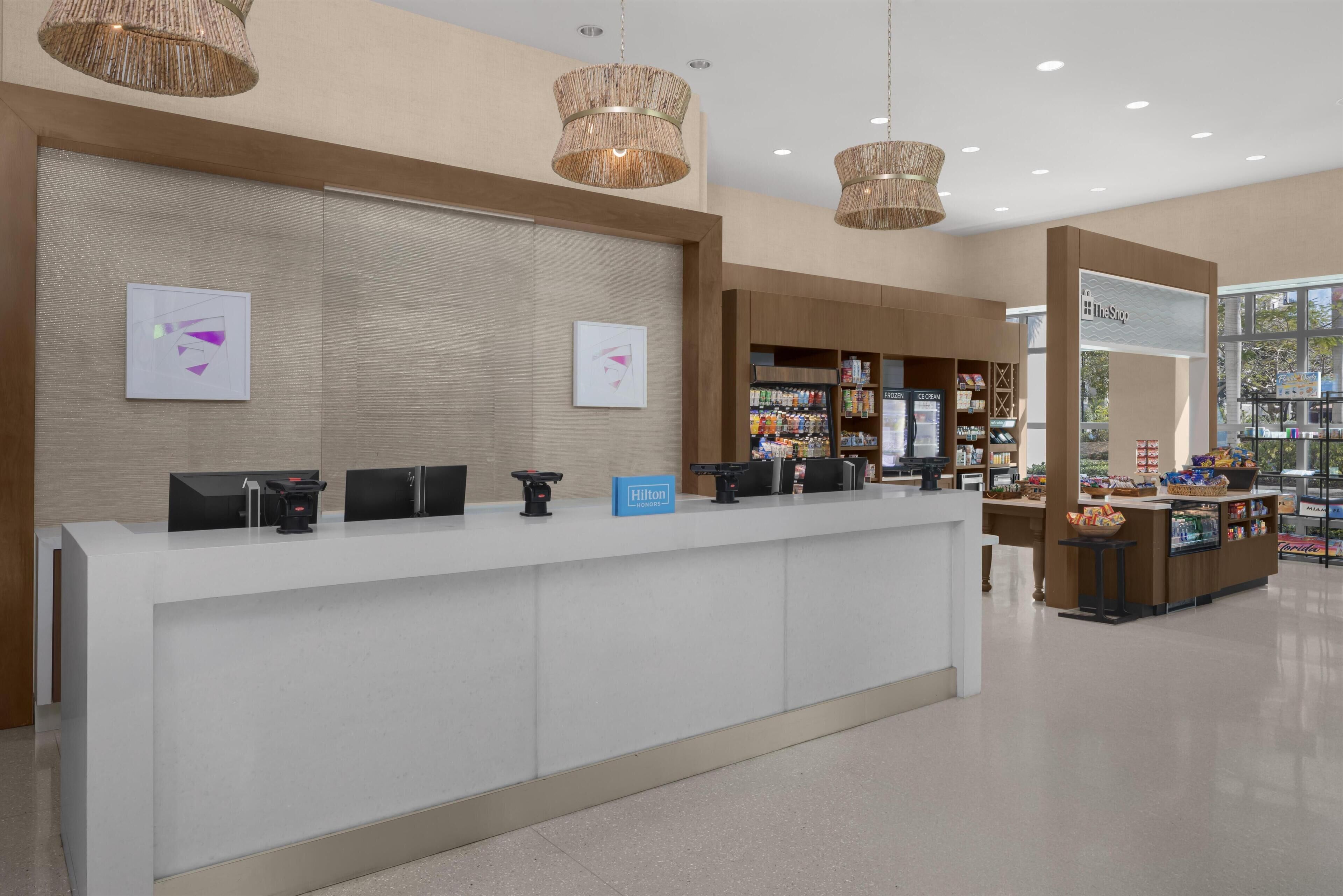 A hotel reception area with a white counter, digital check-in kiosks, a minibar, and a view of the outside.