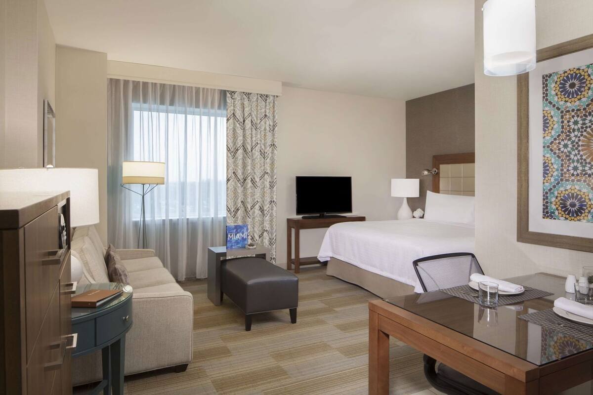 Habitación de hotel con cama, televisor, escritorio, silla, sofá y lámpara.