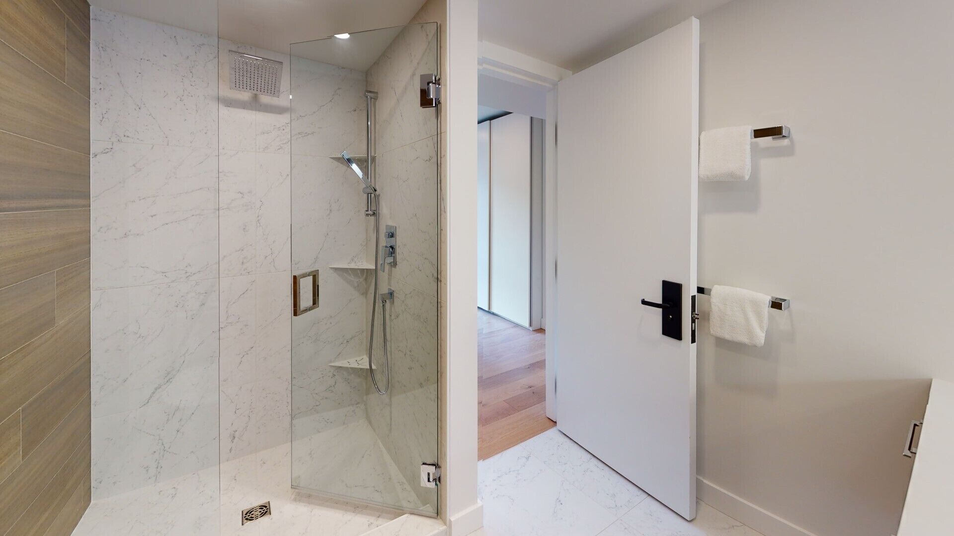 Um banheiro moderno com chuveiro em gabinete de vidro, porta branca com porta-toalhas e piso de madeira.
