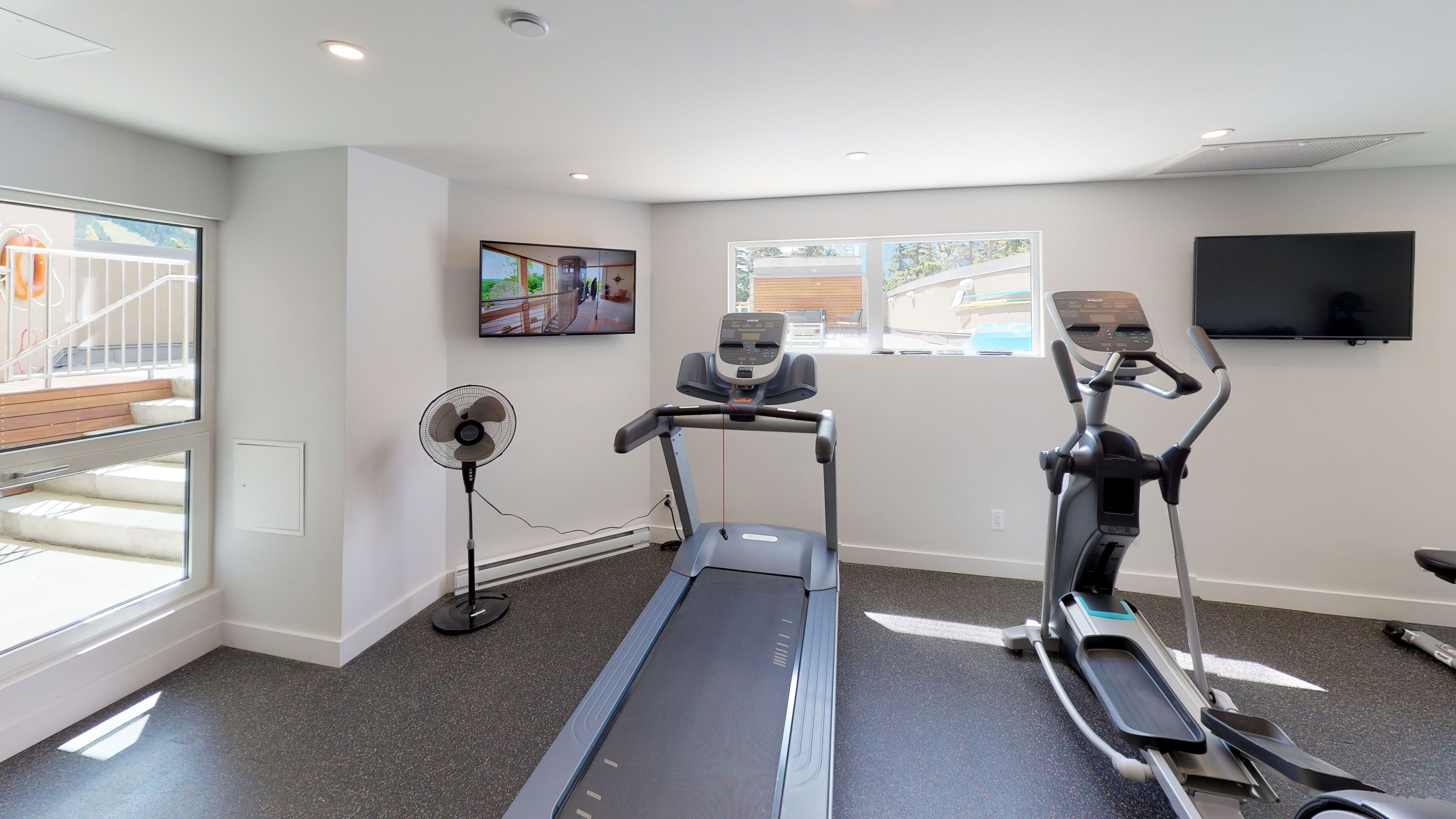 A gym with treadmills, a stationary bike, and a fan. There are windows and a TV on the wall.