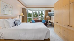 A hotel room with a large bed, a desk, a TV, and a view of trees through the windows.