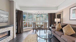 A modern living room with a fireplace, a glass coffee table, a sofa with cushions, a dining area with chairs, and large windows offering a view of buildings.