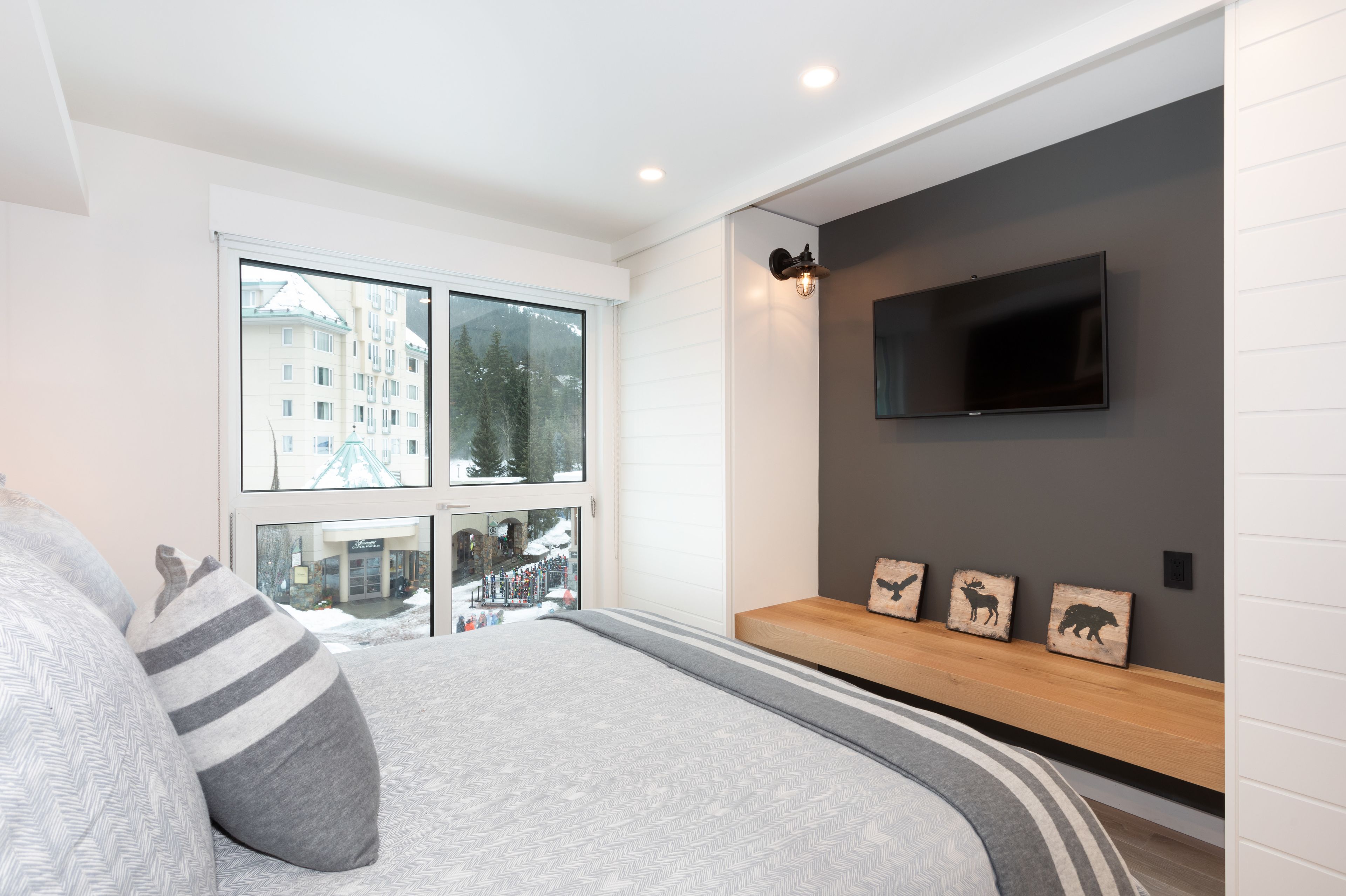Une chambre moderne avec un grand lit, un téléviseur fixé au mur et une fenêtre donnant sur des immeubles et des arbres.