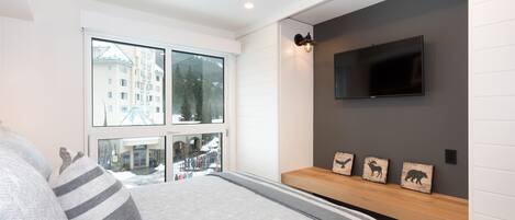 Une chambre moderne avec un grand lit, un téléviseur fixé au mur et une fenêtre donnant sur des immeubles et des arbres.