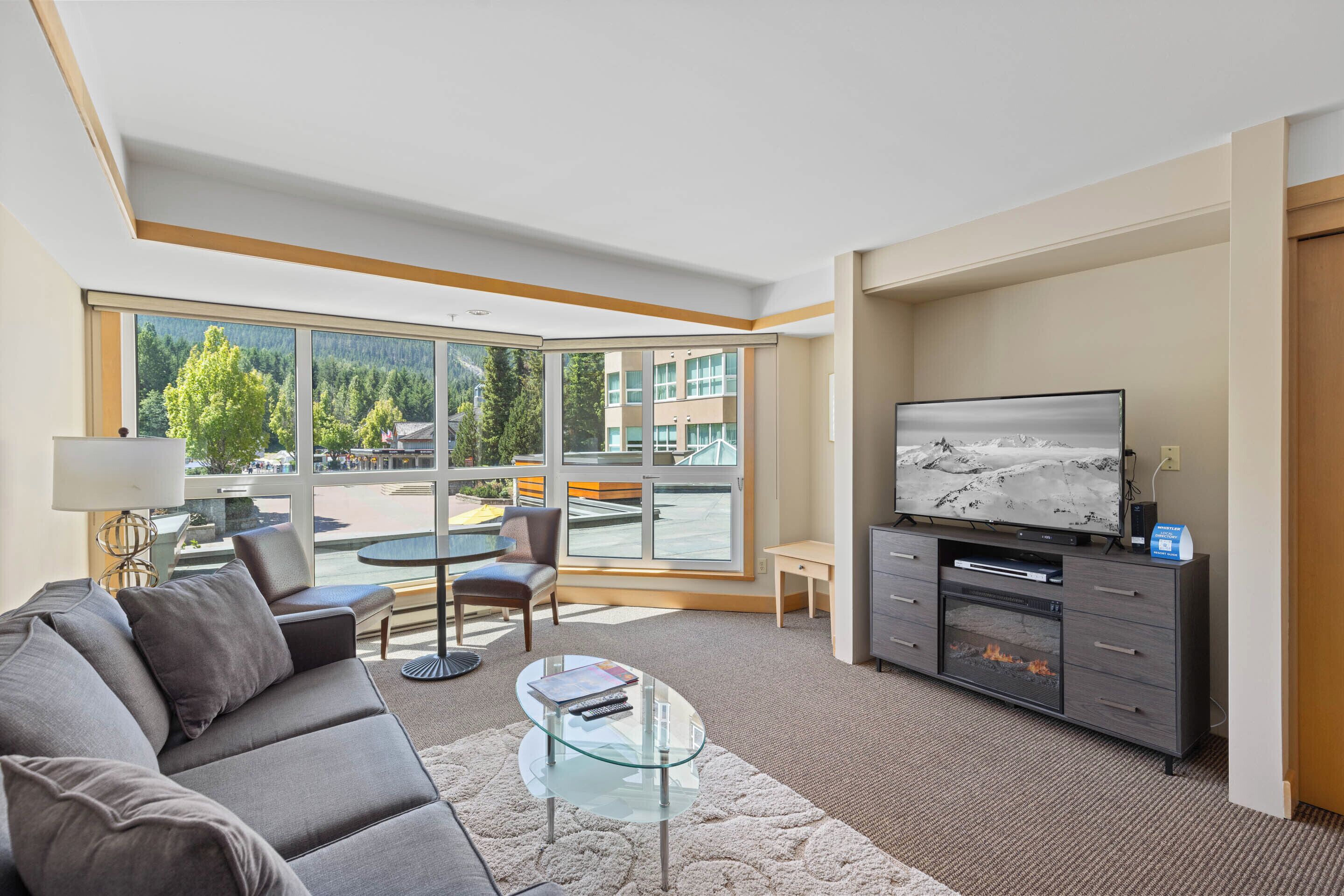 A modern living room with a sofa, coffee table, TV, fireplace, and large windows offering a view of a building and trees.
