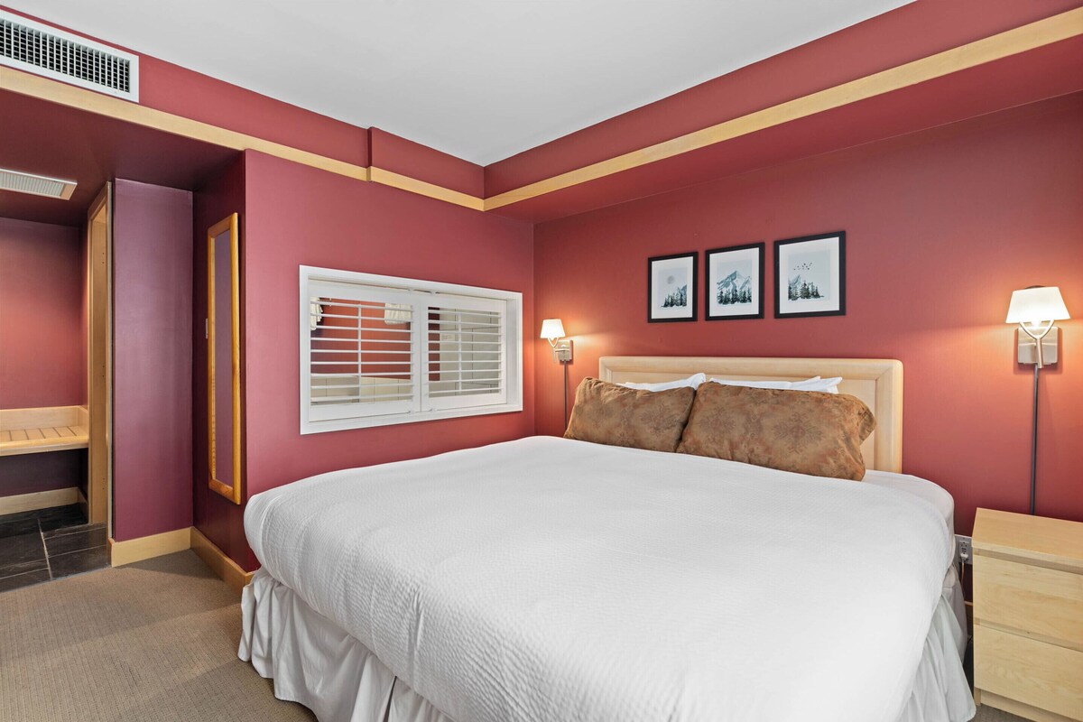 A bedroom with a large bed, a window with blinds, a nightstand, and wall-mounted lamps.