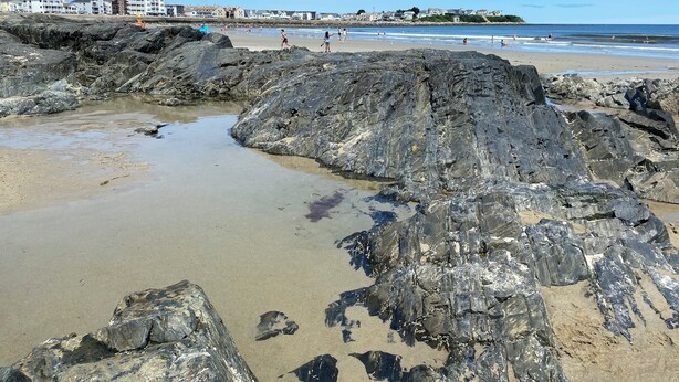 A rocky beach with clear water, a sandy shore, and buildings in the background.
