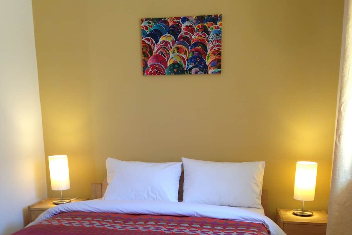 Ein Hotelzimmer mit einem Bett, zwei Nachttischlampen, einem farbenfrohen Bild an der Wand und einer gemusterten Tagesdecke.