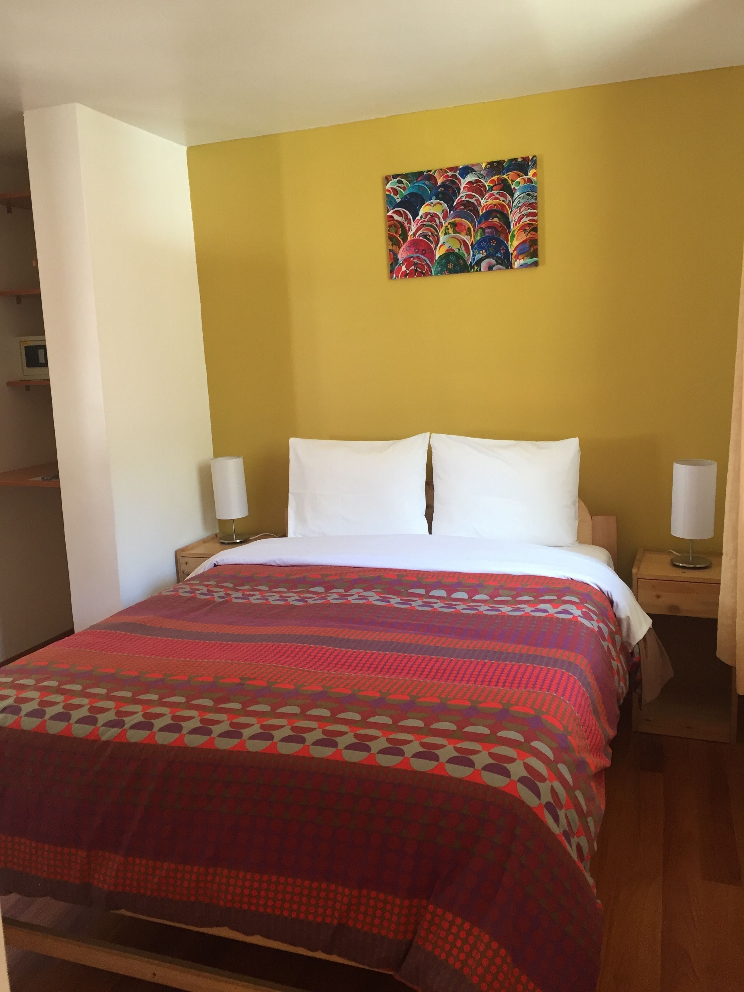 Quarto de hotel com uma cama, duas mesas de cabeceira, um quadro pendurado na parede e piso de madeira.
