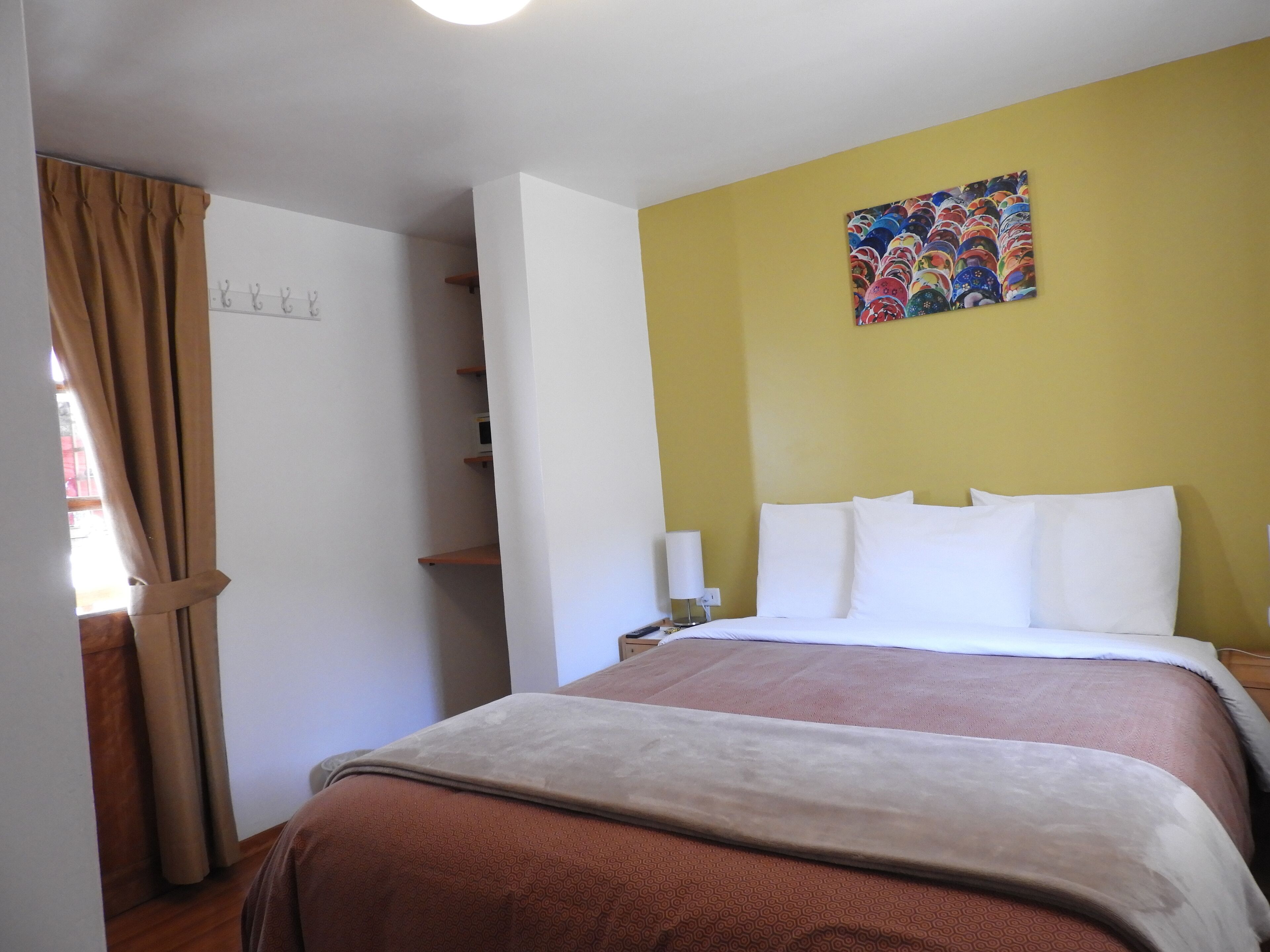 A hotel room with a bed, a nightstand, a lamp, a wall with a colorful artwork, and a door with a curtain.