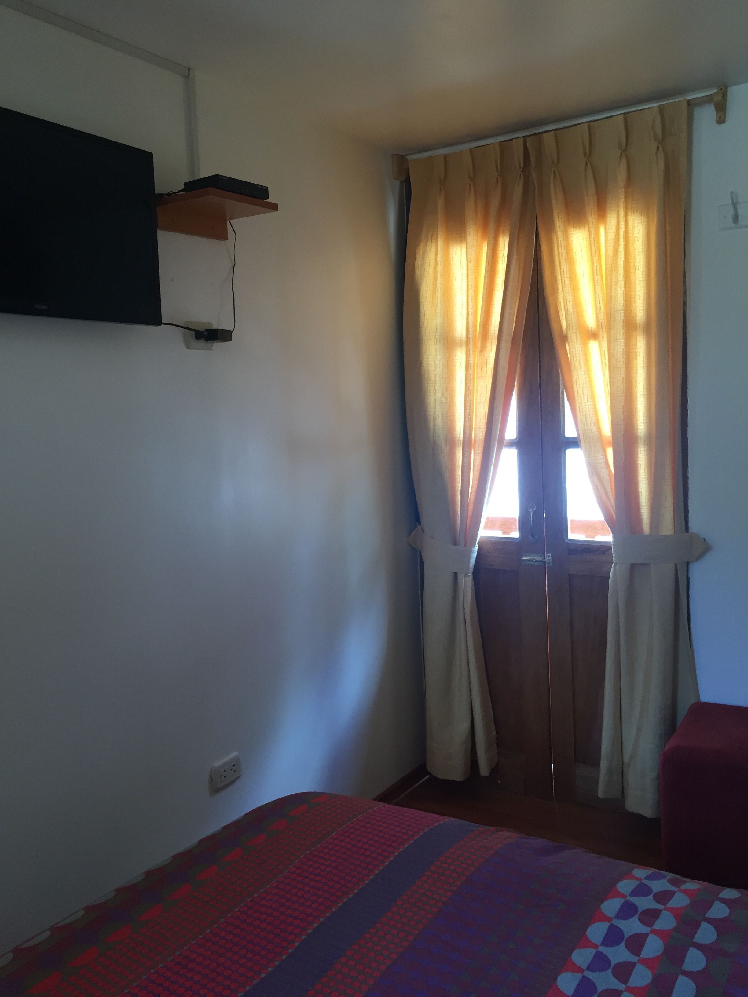 A room with a bed, a window with curtains, a wall-mounted shelf, and a red chair.