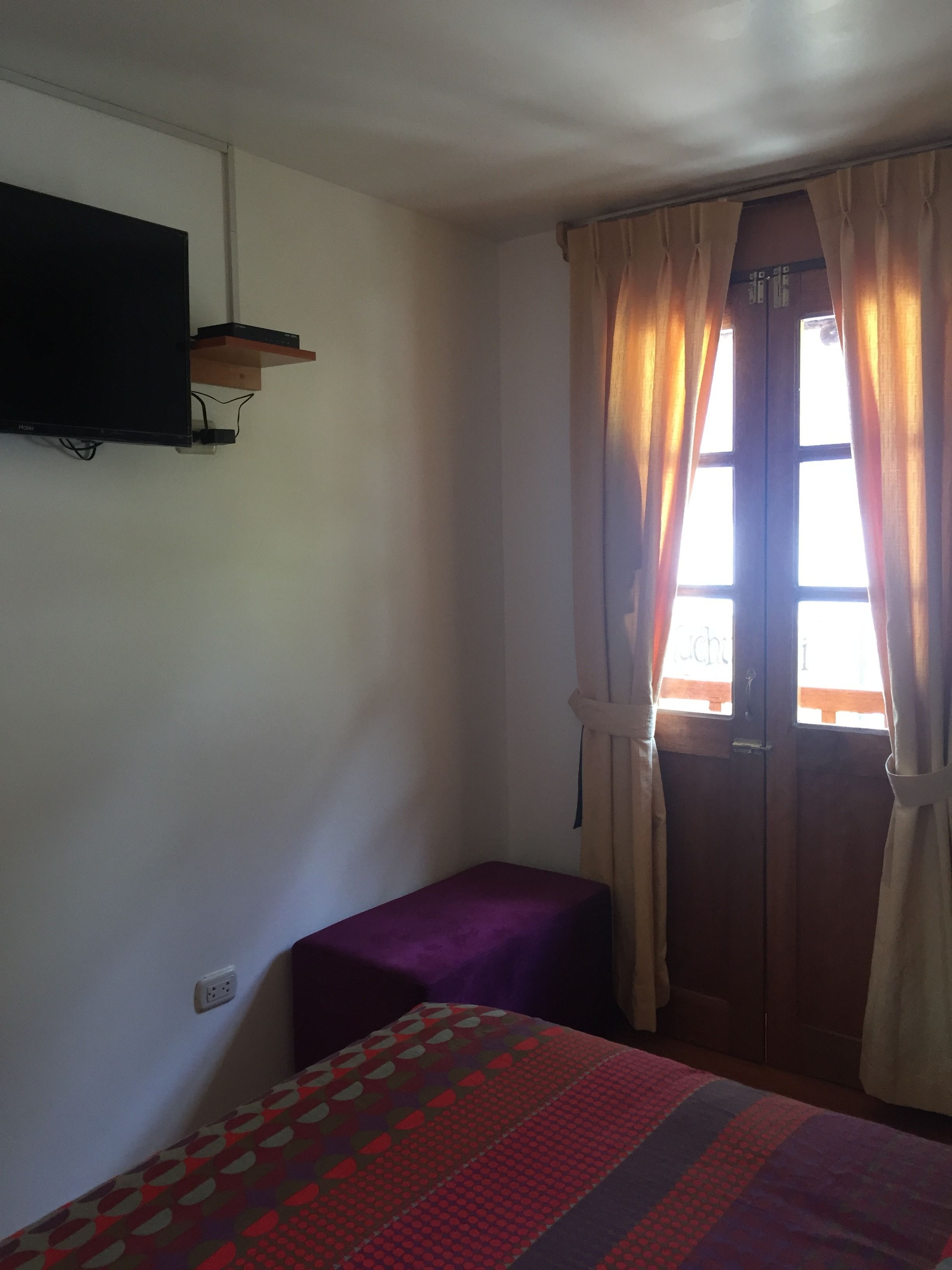 A small room with a bed, a TV mounted on the wall, and a window with curtains.
