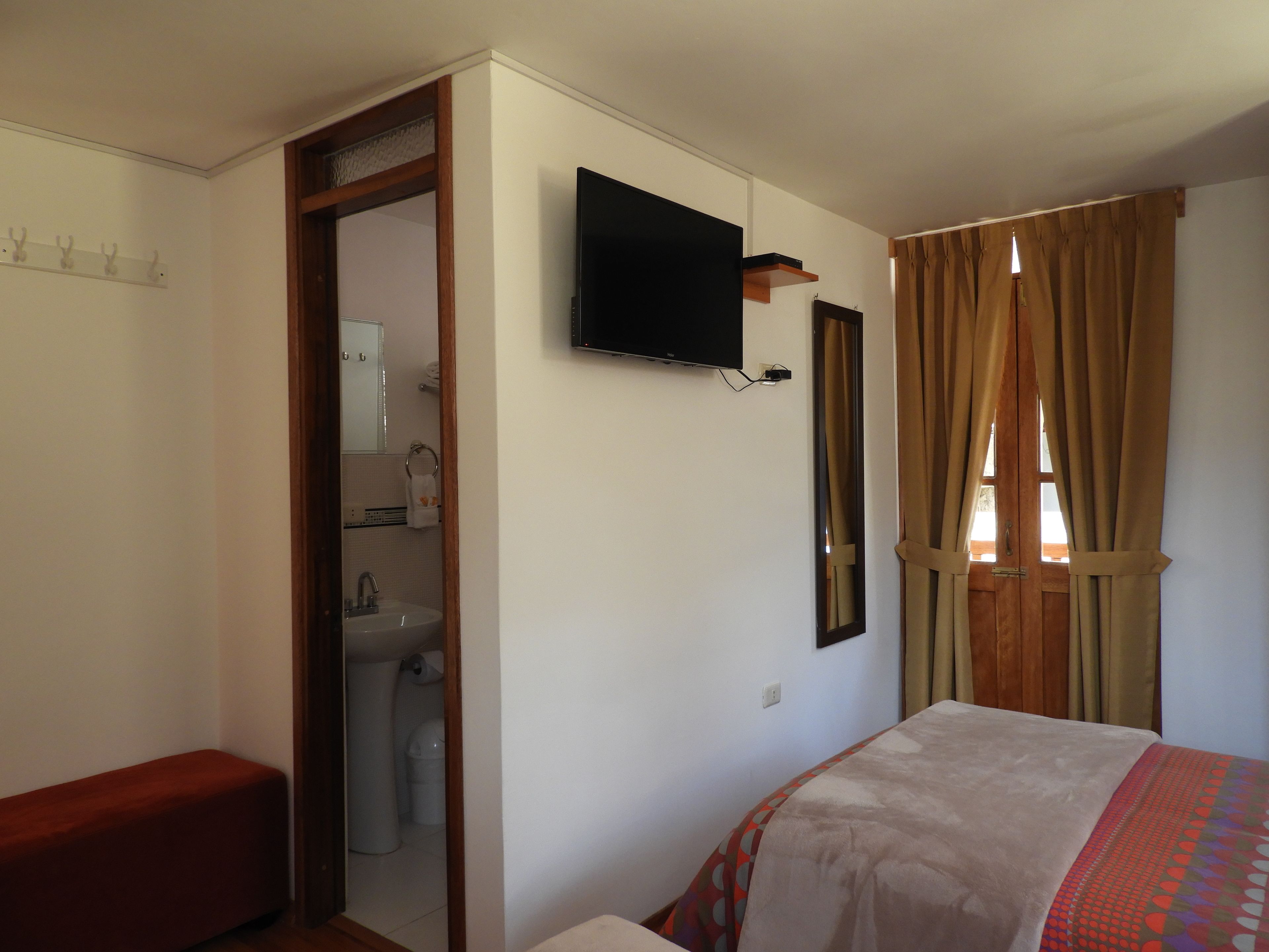 Een slaapkamer met een bed, een aan de muur bevestigde televisie, een zichtbare badkamer door een open deur en een raam met gordijnen.