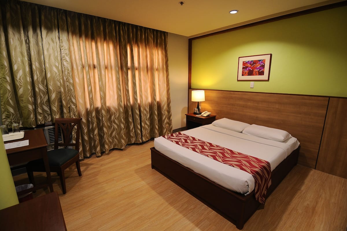 A hotel room with a large bed, a desk with a chair, a nightstand with a lamp, and a painting on the wall.