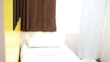 A small, single-bed room with a white mattress, brown curtains, and a white window blind.