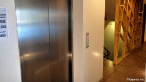 Ein moderner Aufzug mit Metalloptik, einem wandmontierten Notrufknopf und einer Holzwand mit geometrischem Muster.
