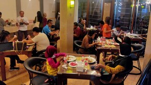 A restaurant with multiple tables and chairs, people dining, and a view of the street outside. - Hotel Grand Sumatera (Surabaya)