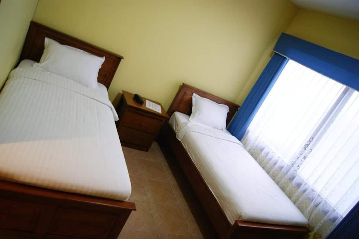 Habitación de hotel con dos camas, cabecero de madera, mesita de noche con teléfono y ventana con cortinas.